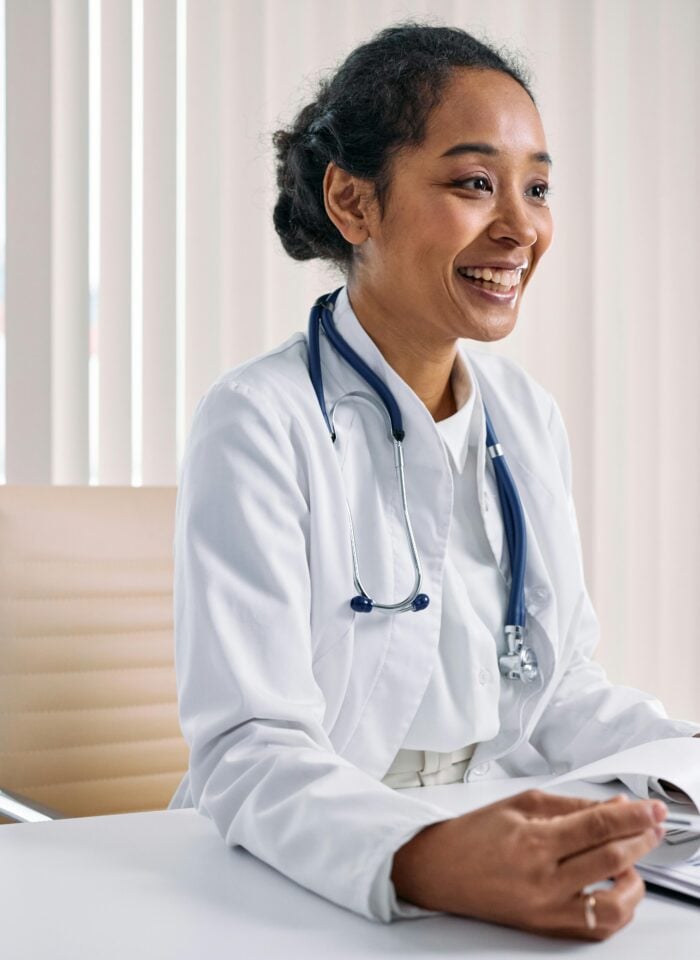 Cheerful female doctor wearing a white coat and stethoscope sitting at a desk during a consultation.