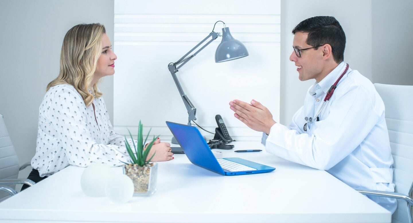 Male doctor in a white coat speaking with a female patient across a desk with a laptop and lamp in a bright office.