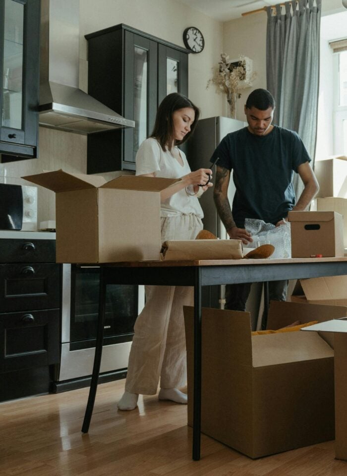Family unpacking moving boxes in a kitchen while a child plays inside a box.
