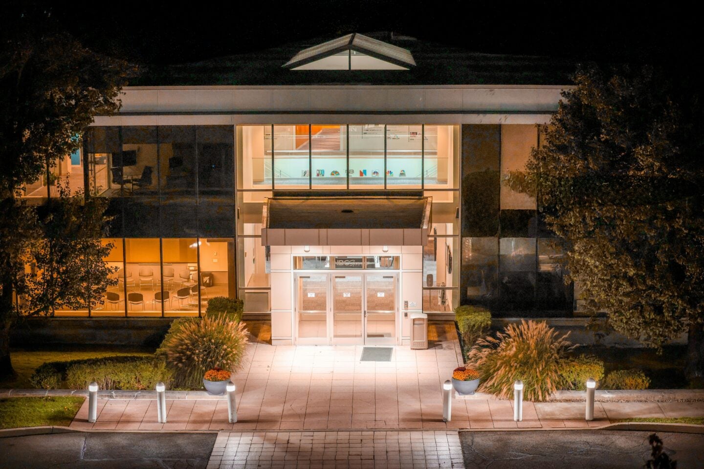 Illuminated office building entrance at night with glass doors and landscaped surroundings.