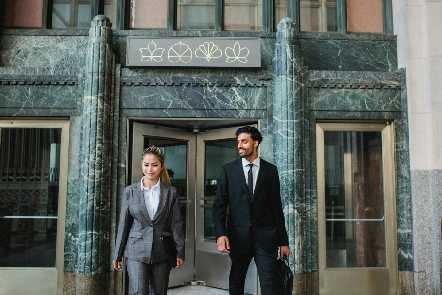 Two business professionals in suits walking out of an office building entrance.