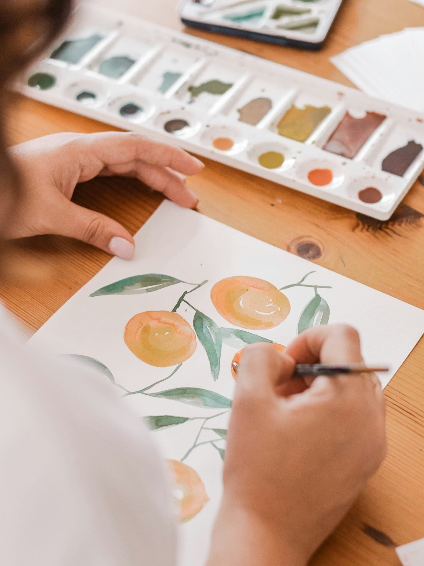 Person painting orange fruit and green leaves with watercolors on white paper at a wooden table.