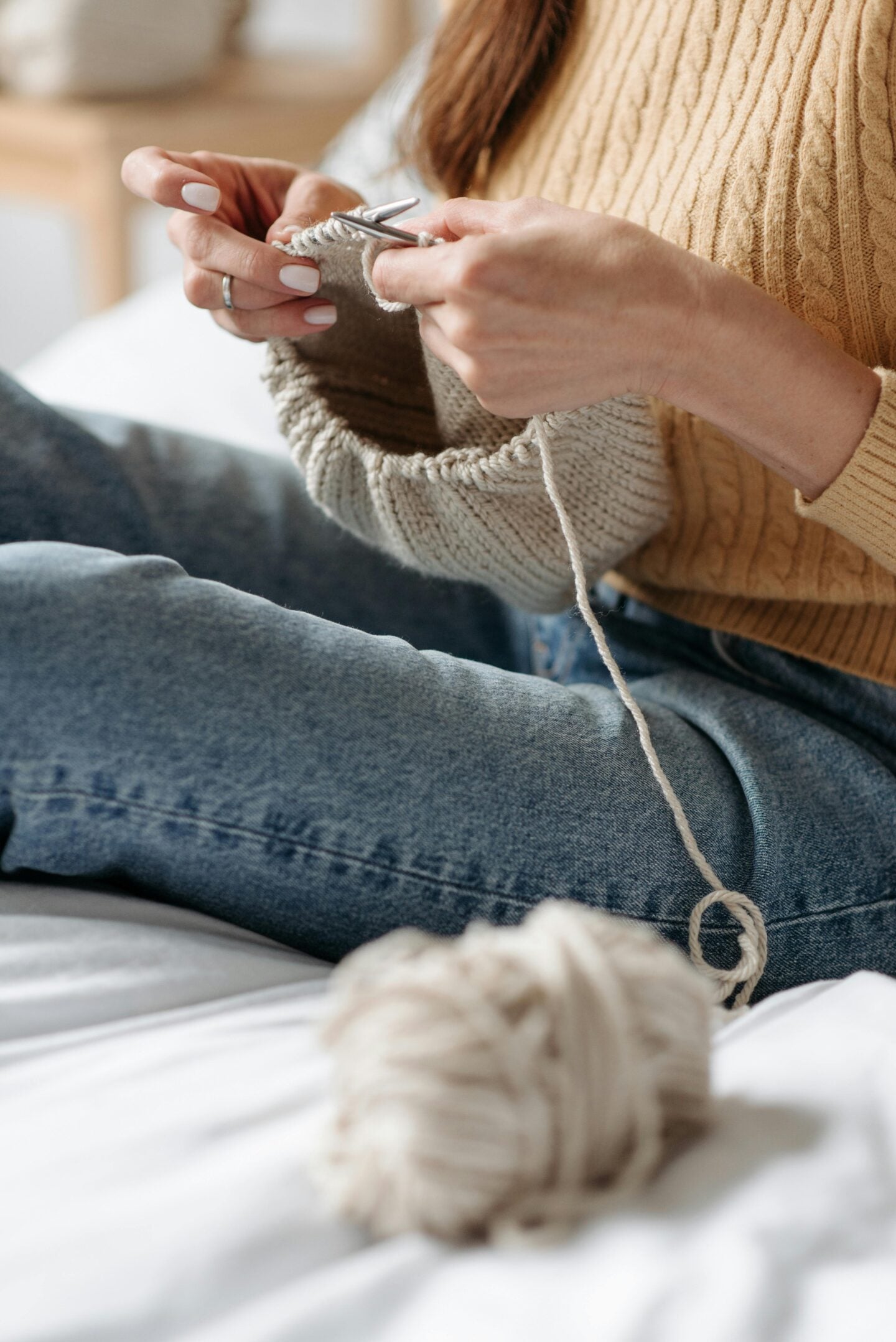 Woman knitting a beige piece of fabric while sitting comfortably on a bed with yarn nearby.