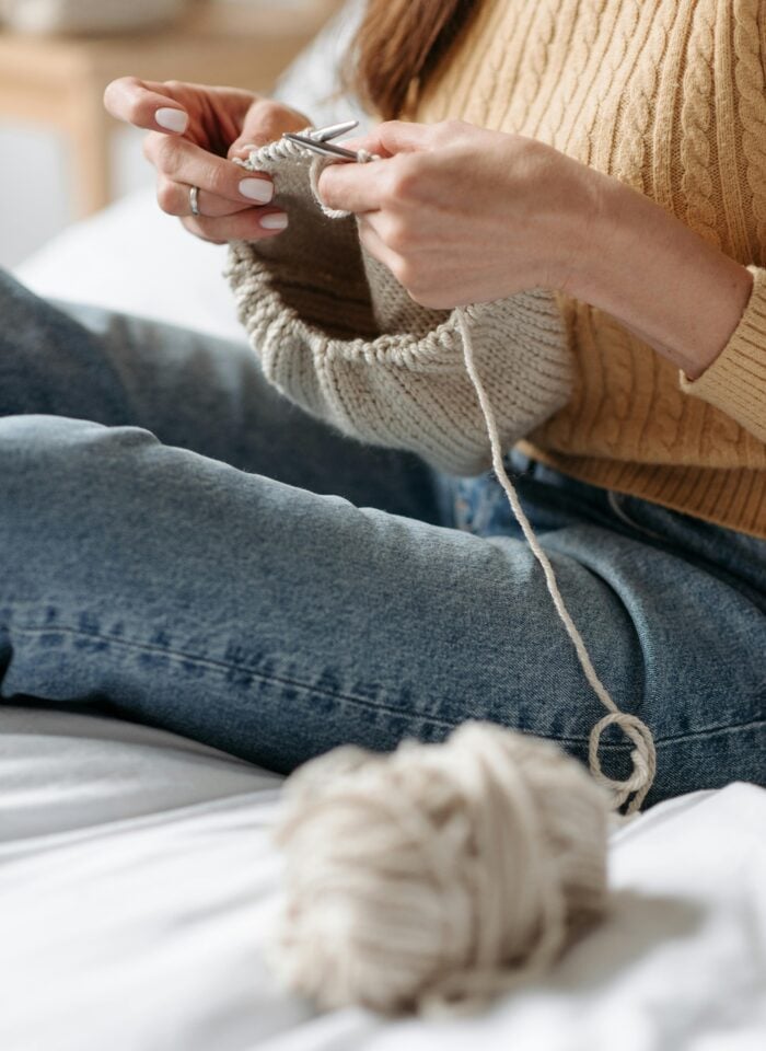 Woman knitting a beige piece of fabric while sitting comfortably on a bed with yarn nearby.
