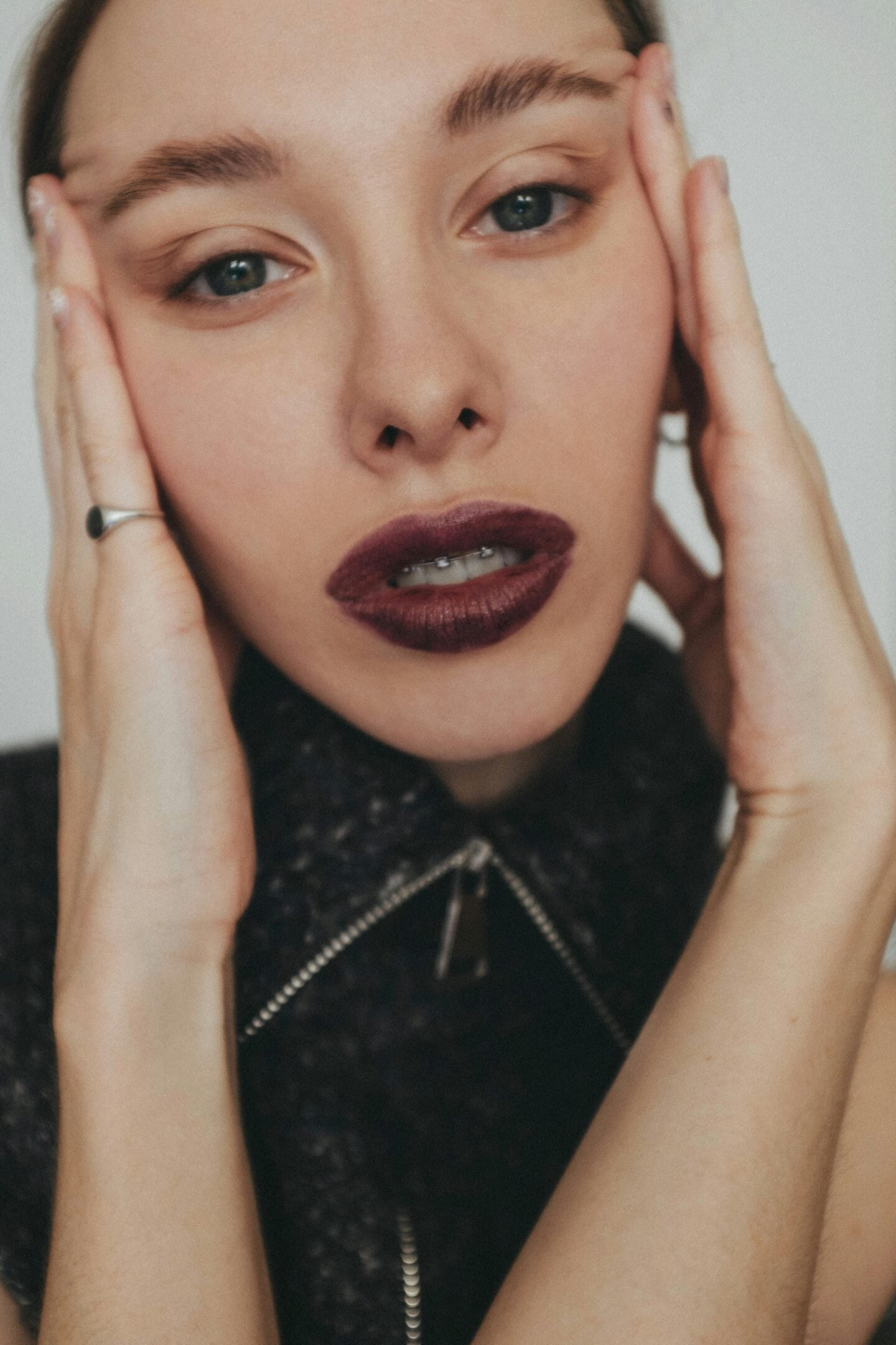 Portrait of a young woman with dark plum lipstick and subtle makeup, revealing clear braces on her upper teeth.
