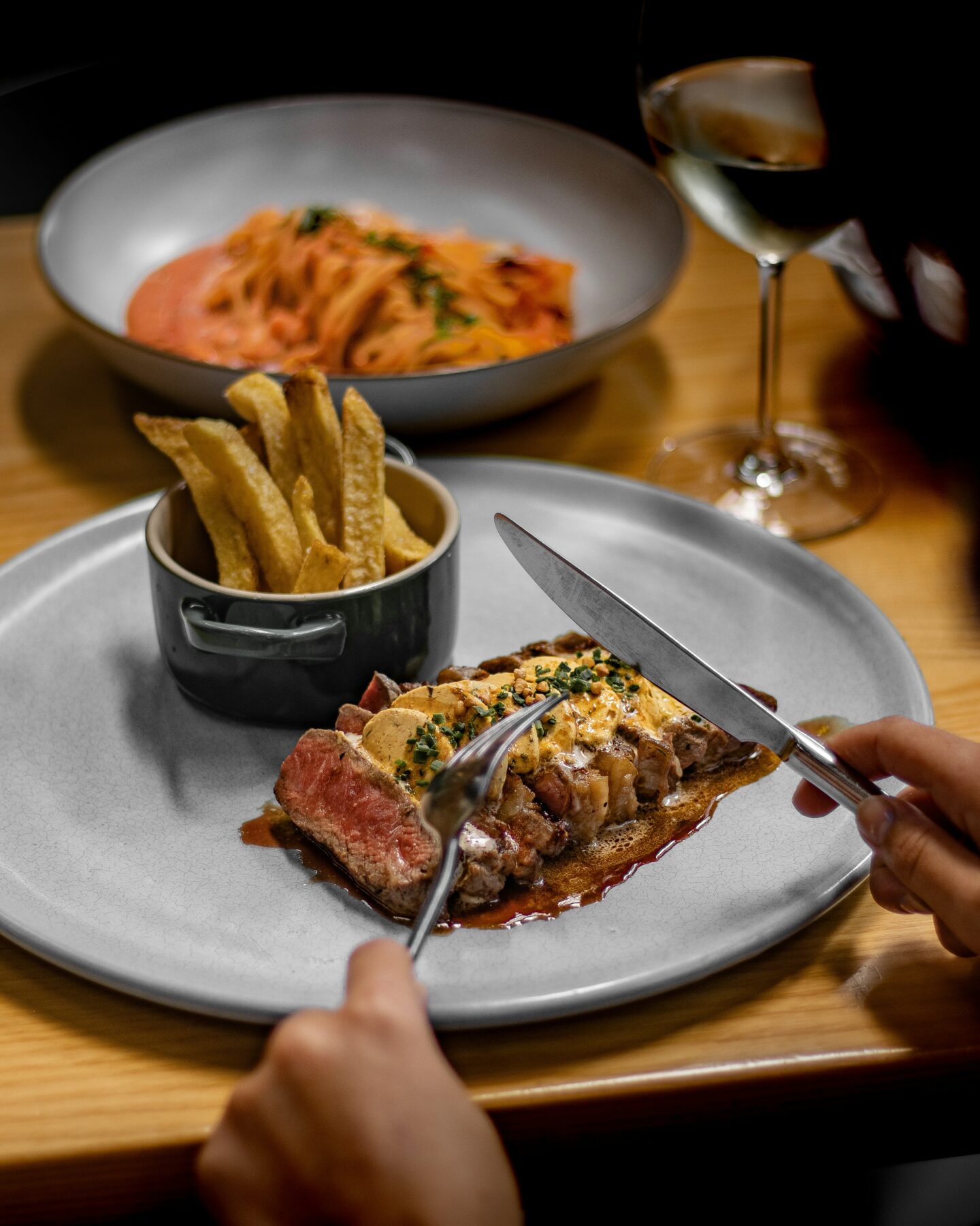 Steakhouse meal with steak, fries, and pasta served alongside a glass of wine.