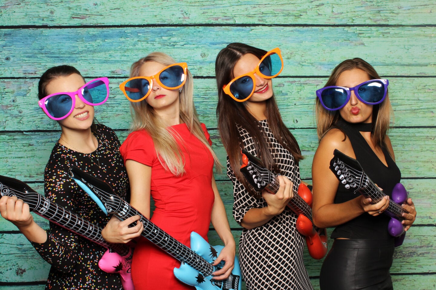 Group of women wearing oversized novelty sunglasses and holding inflatable guitars posing for a photo at at party photobooth.