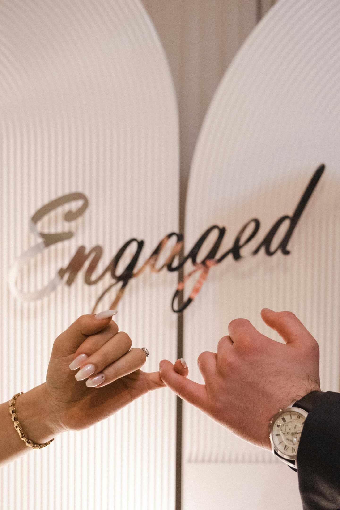 Couple linking pinky fingers in front of a decorative sign that says 'Engaged.'