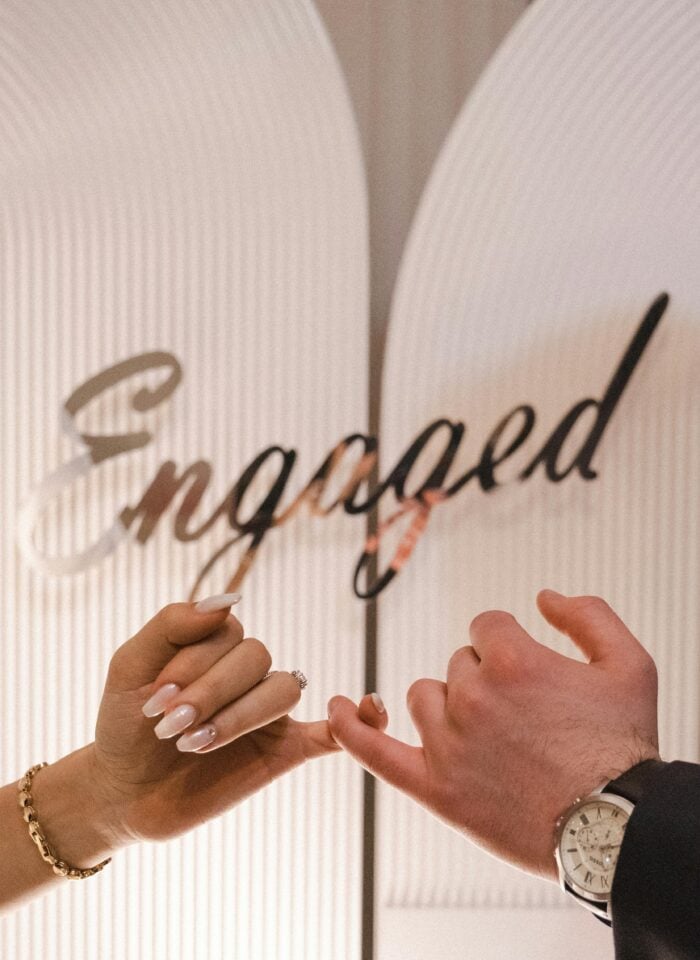 Couple linking pinky fingers in front of a decorative sign that says 'Engaged.'