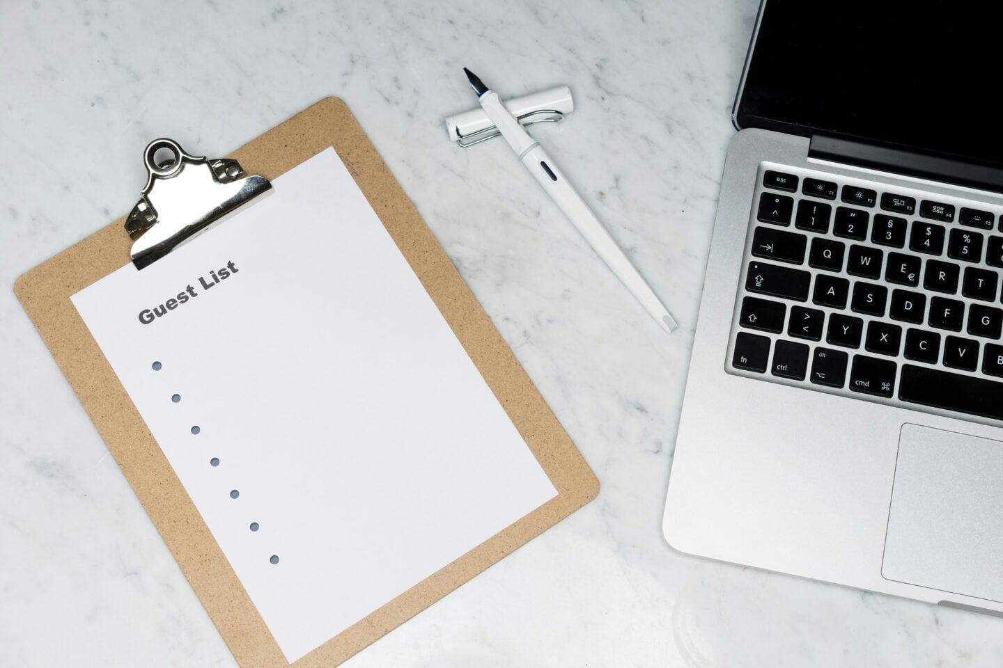 Clipboard with a blank guest list paper beside a pen and laptop on a marble surface.