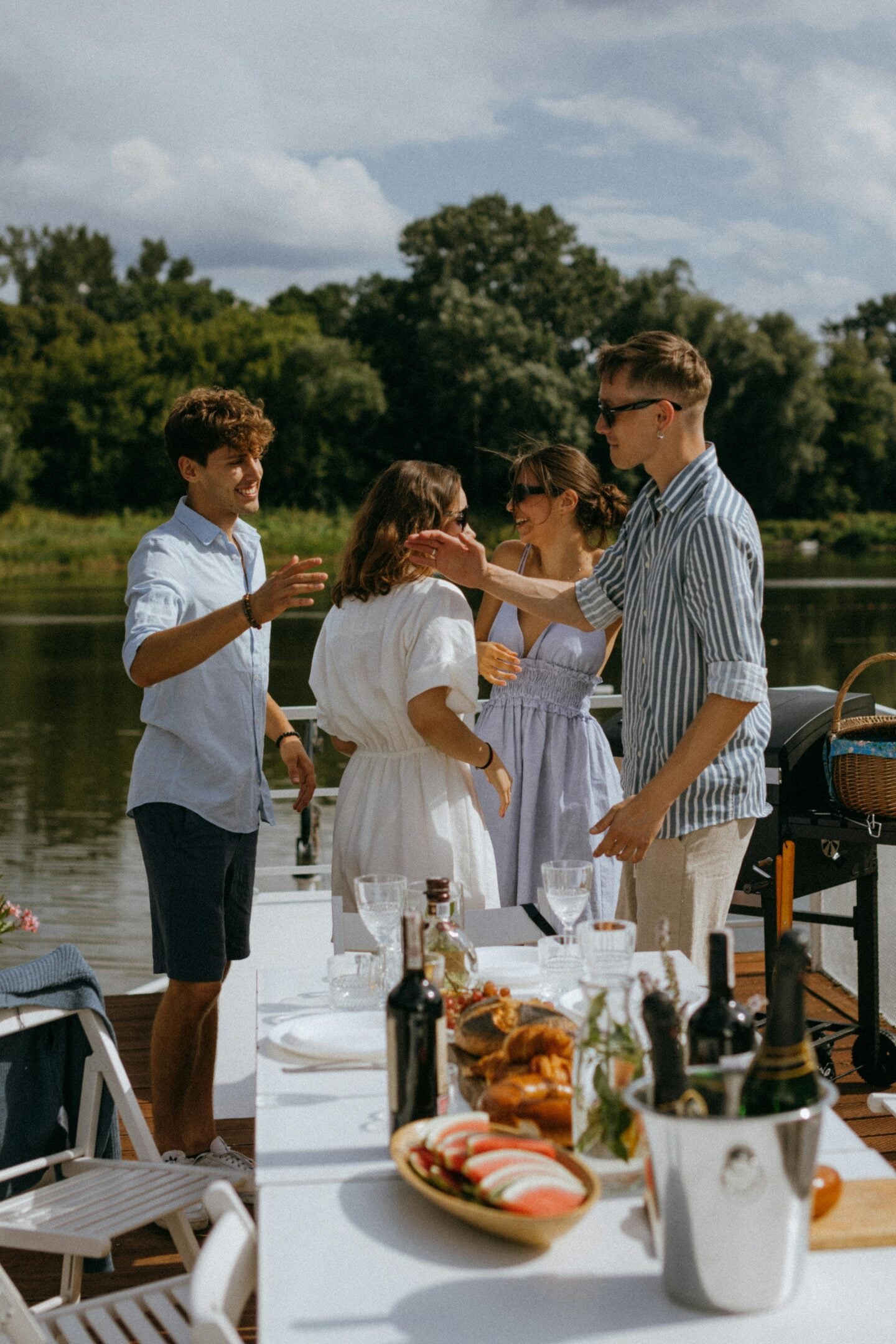 Friends greeting and hugging at an outdoor riverside gathering with food and drinks on the table.