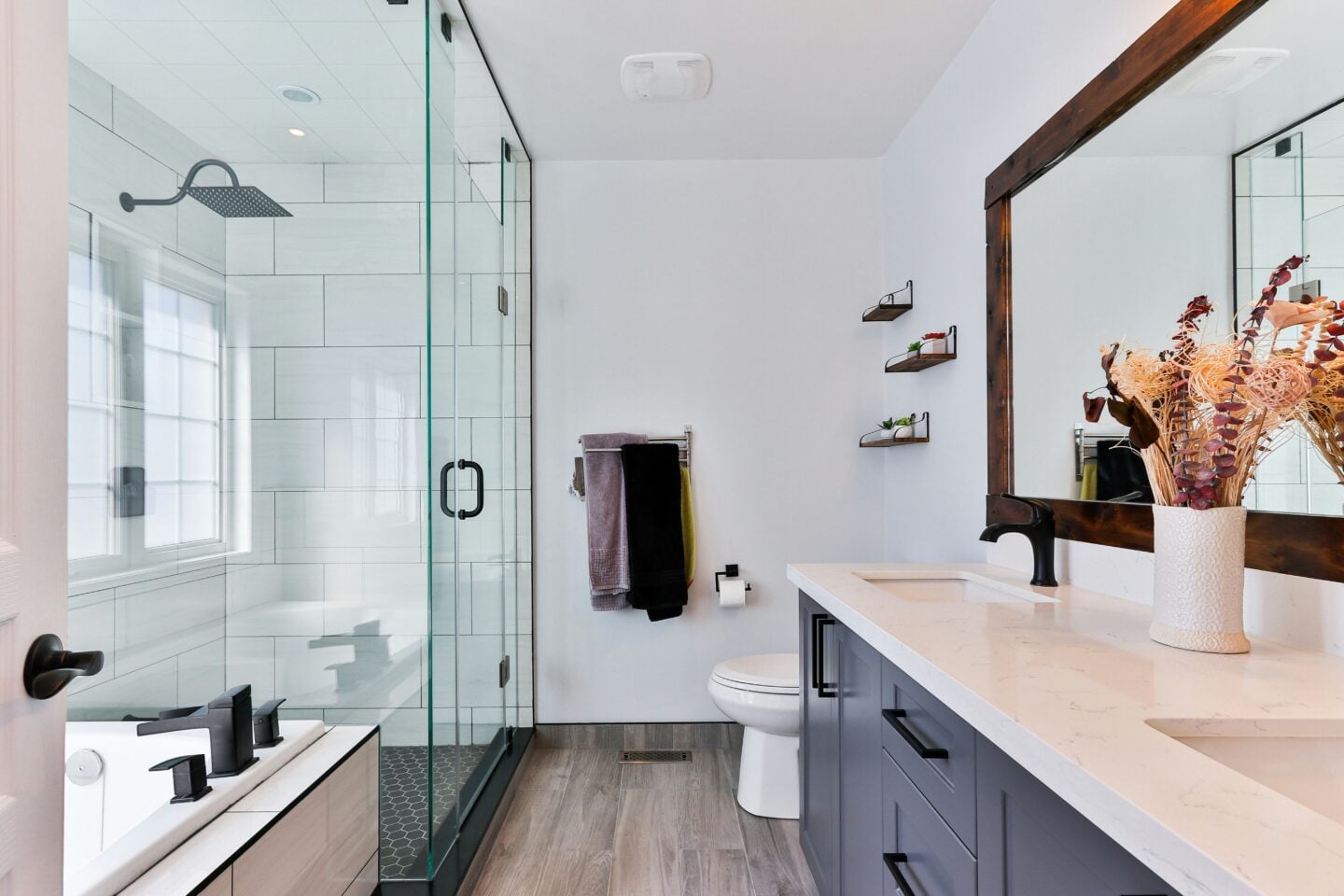 Bright contemporary bathroom with glass shower, double vanity, black fixtures, and wood-framed mirror over neutral countertops.