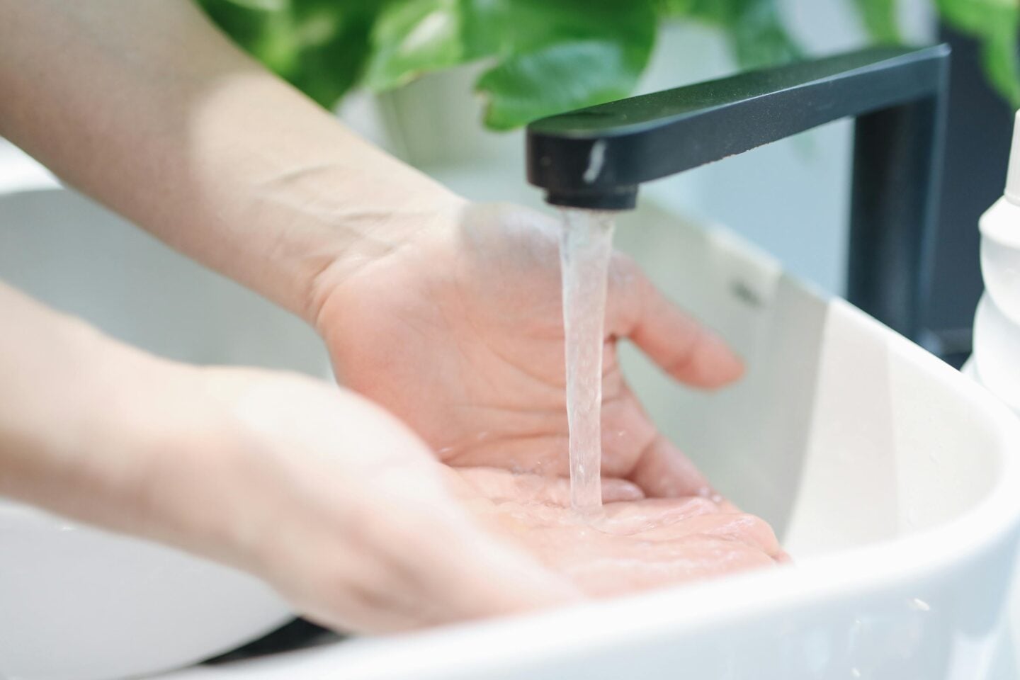 Close-up of hands being washed under a modern black touchless faucet with a white sink and green plant background.