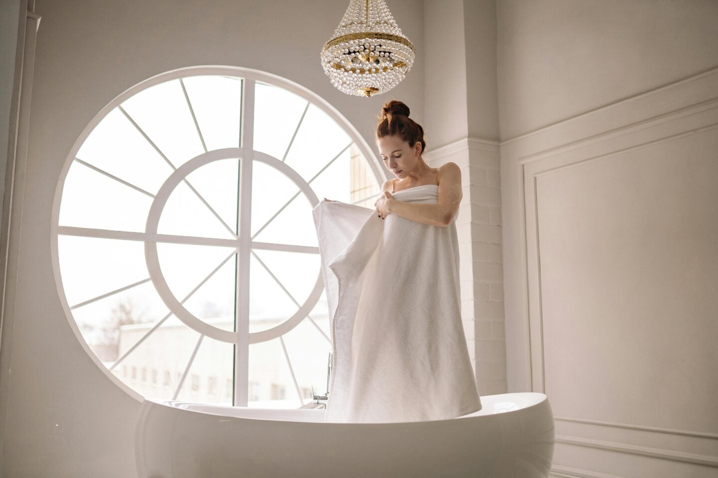 Woman in a white towel standing in a freestanding bathtub beneath a round window and crystal chandelier.