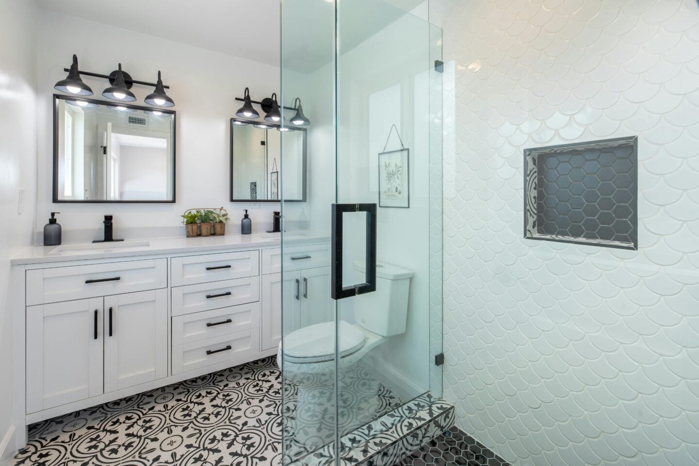 Stylish white bathroom with patterned black-and-white floor tiles, glass shower, and double vanity with black hardware.