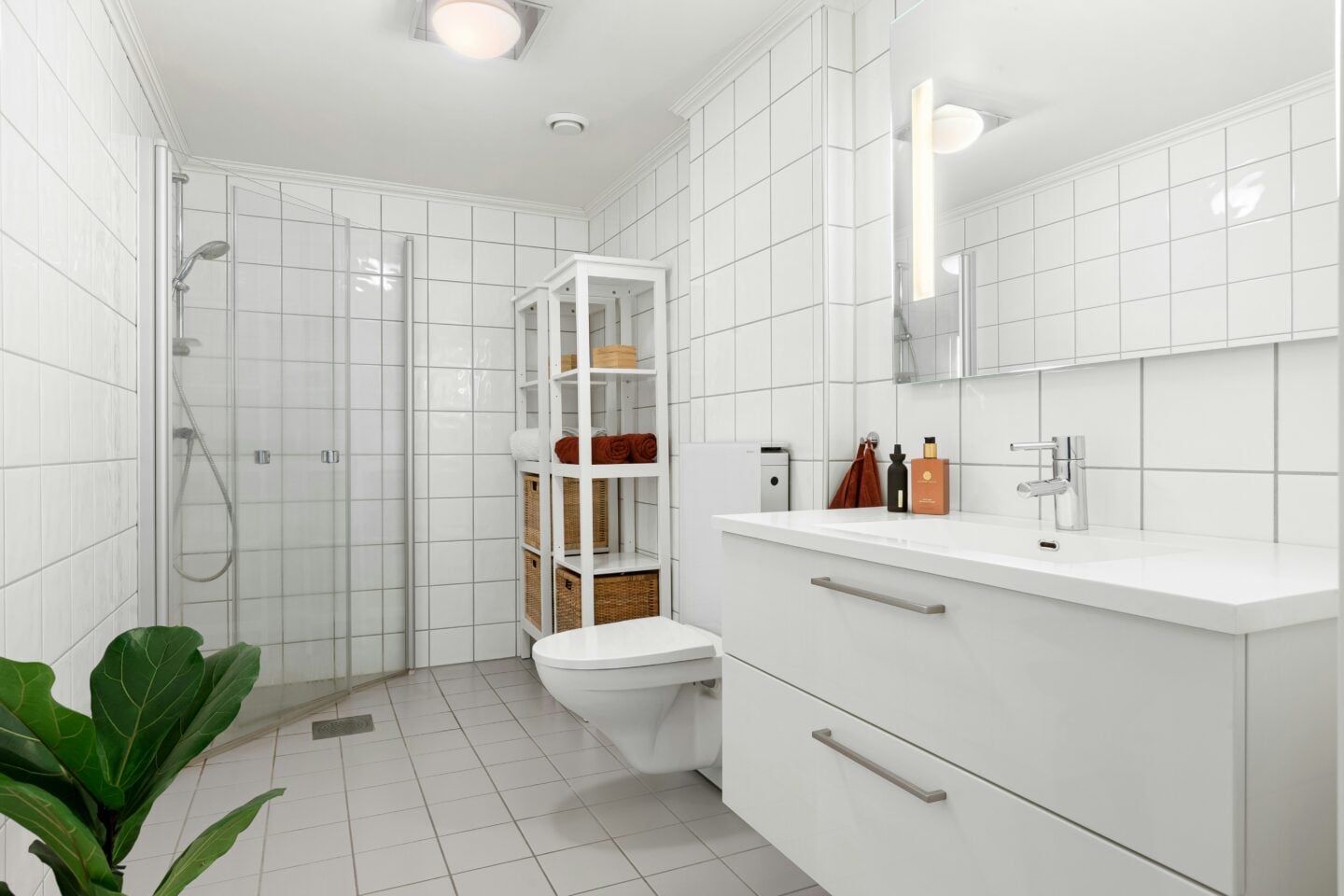 Bright white tiled bathroom with glass shower, floating vanity, open shelving, and warm-toned towels and baskets.