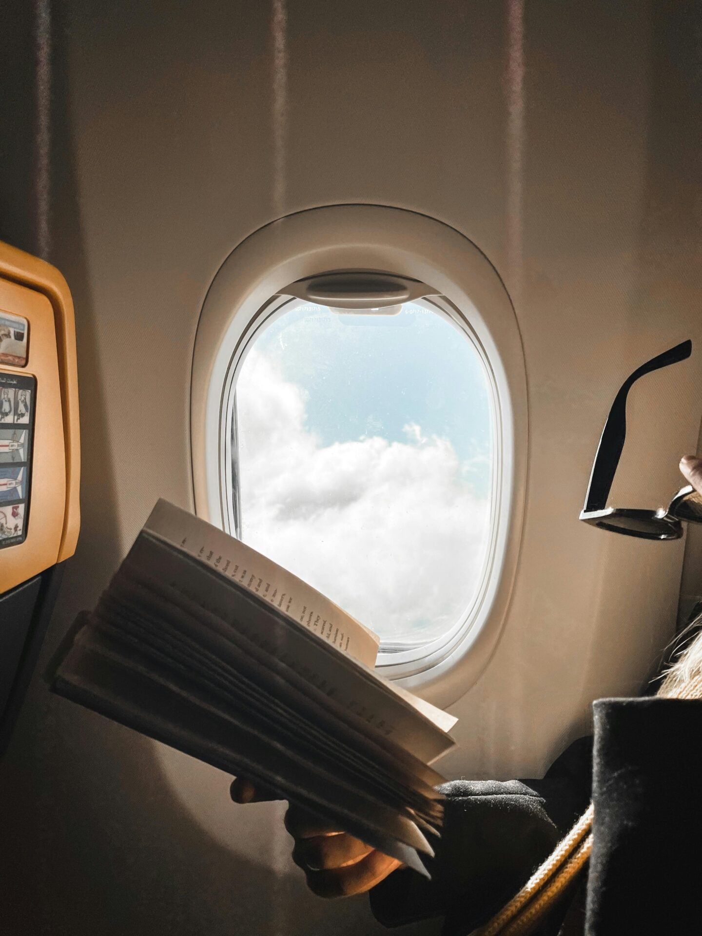 Passenger reading a book beside an airplane window with a view of clouds outside.