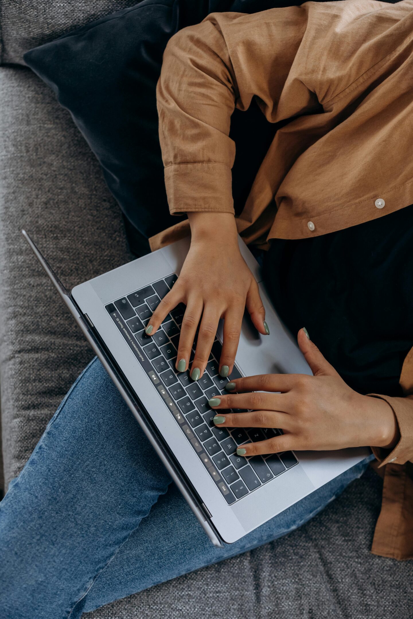 Close-up of a person with green painted nails typing on a laptop keyboard while sitting on a couch.