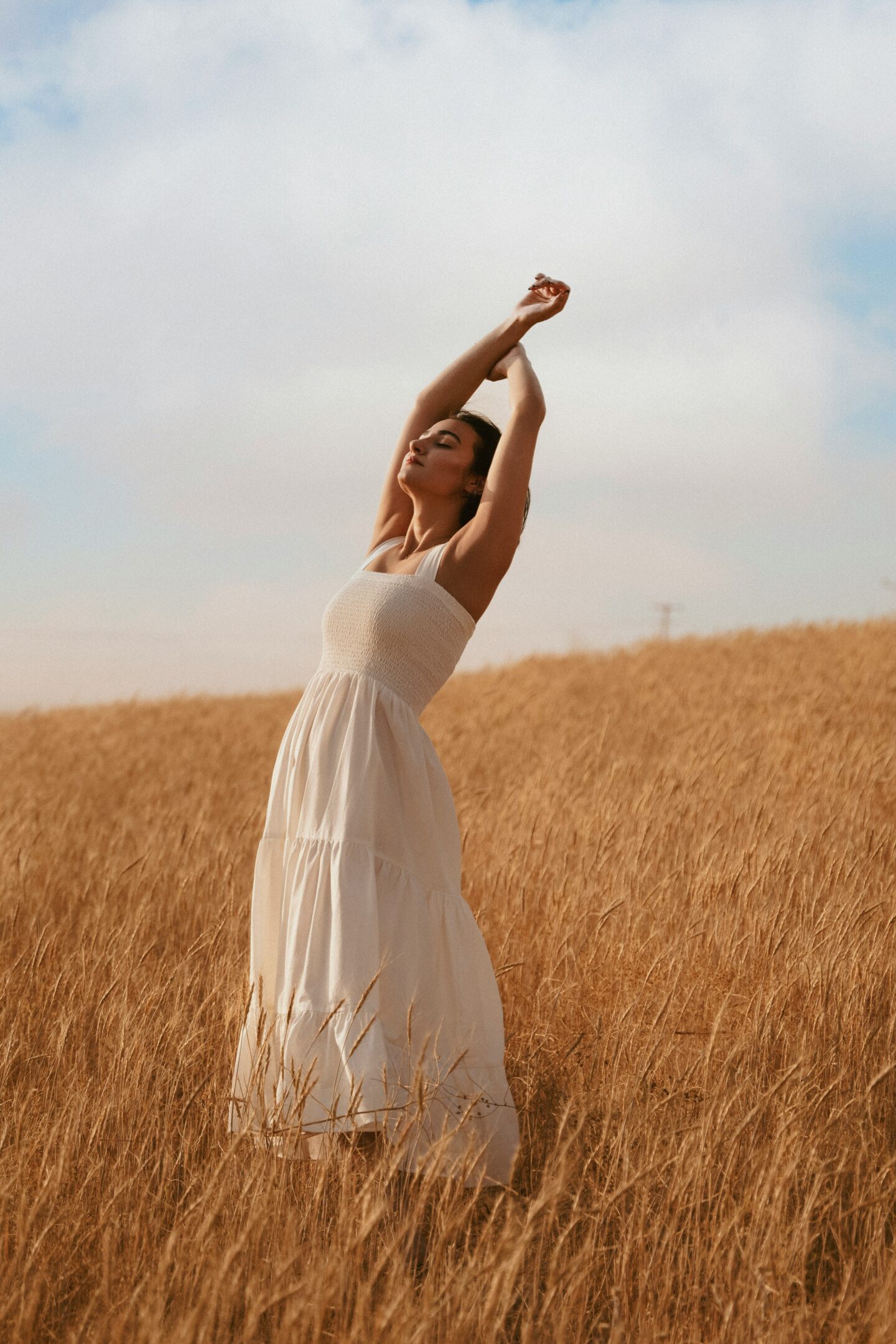 A woman in a flowing white dress stands in a golden wheat field with arms raised, enjoying the open sky.