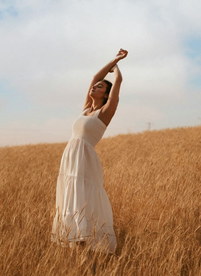 A woman in a flowing white dress stands in a golden wheat field with arms raised, enjoying the open sky.
