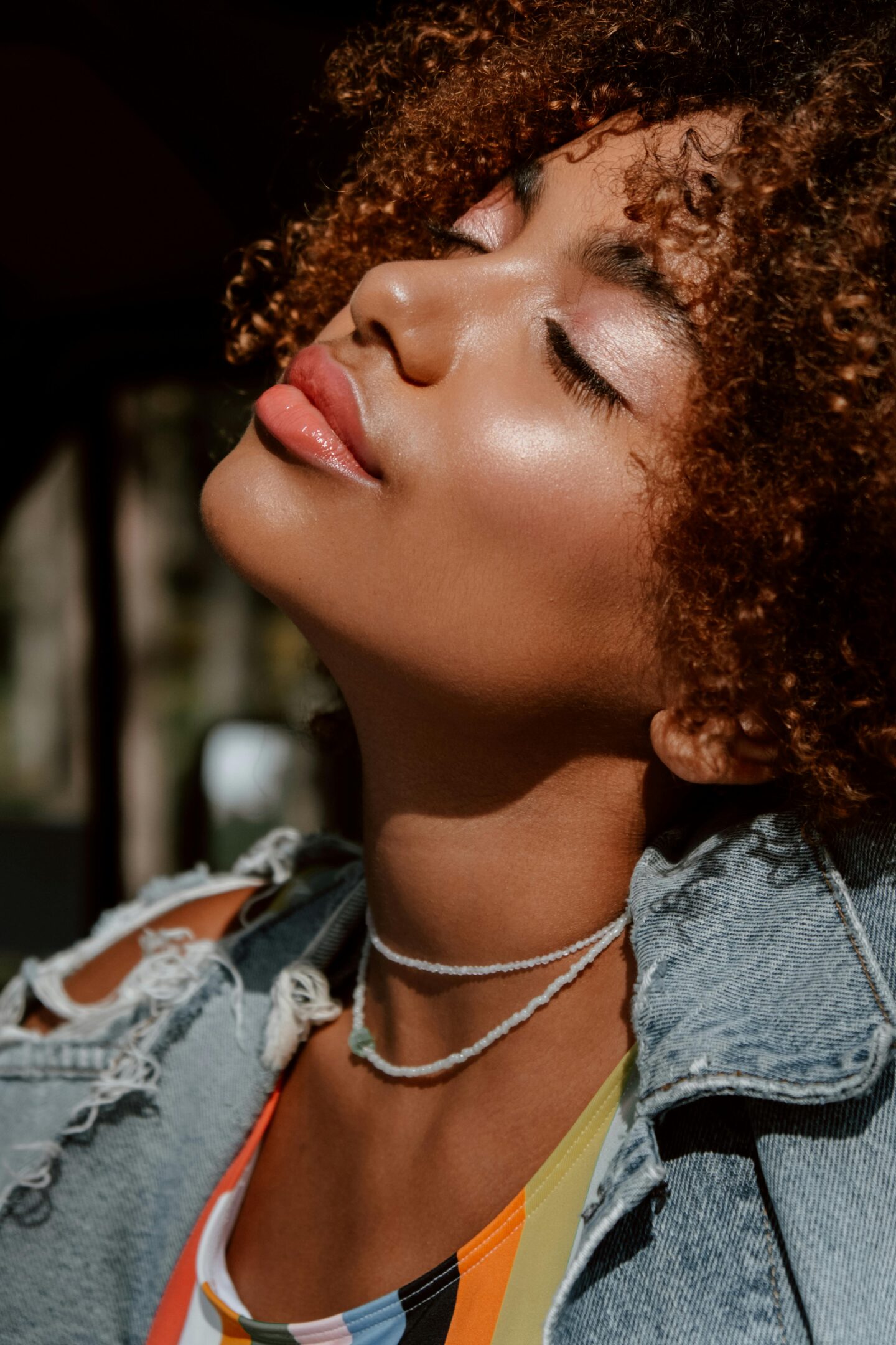 A close-up of a woman with curly hair, glowing skin, and glossy lips tilting her head back in sunlight, wearing a denim jacket and beaded necklaces.