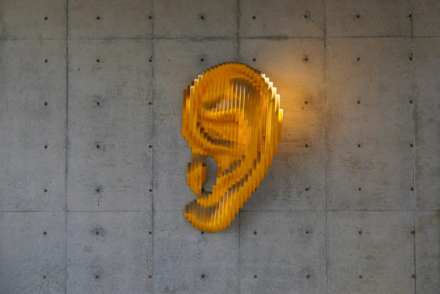 Modern wall art of a large yellow ear sculpture mounted on a gray concrete wall.