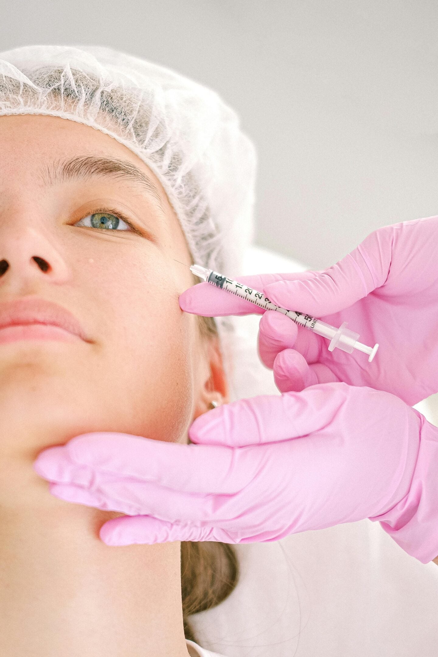 Close-up of a cosmetic procedure where a practitioner wearing pink gloves administers a facial injection to a client in a medical cap.