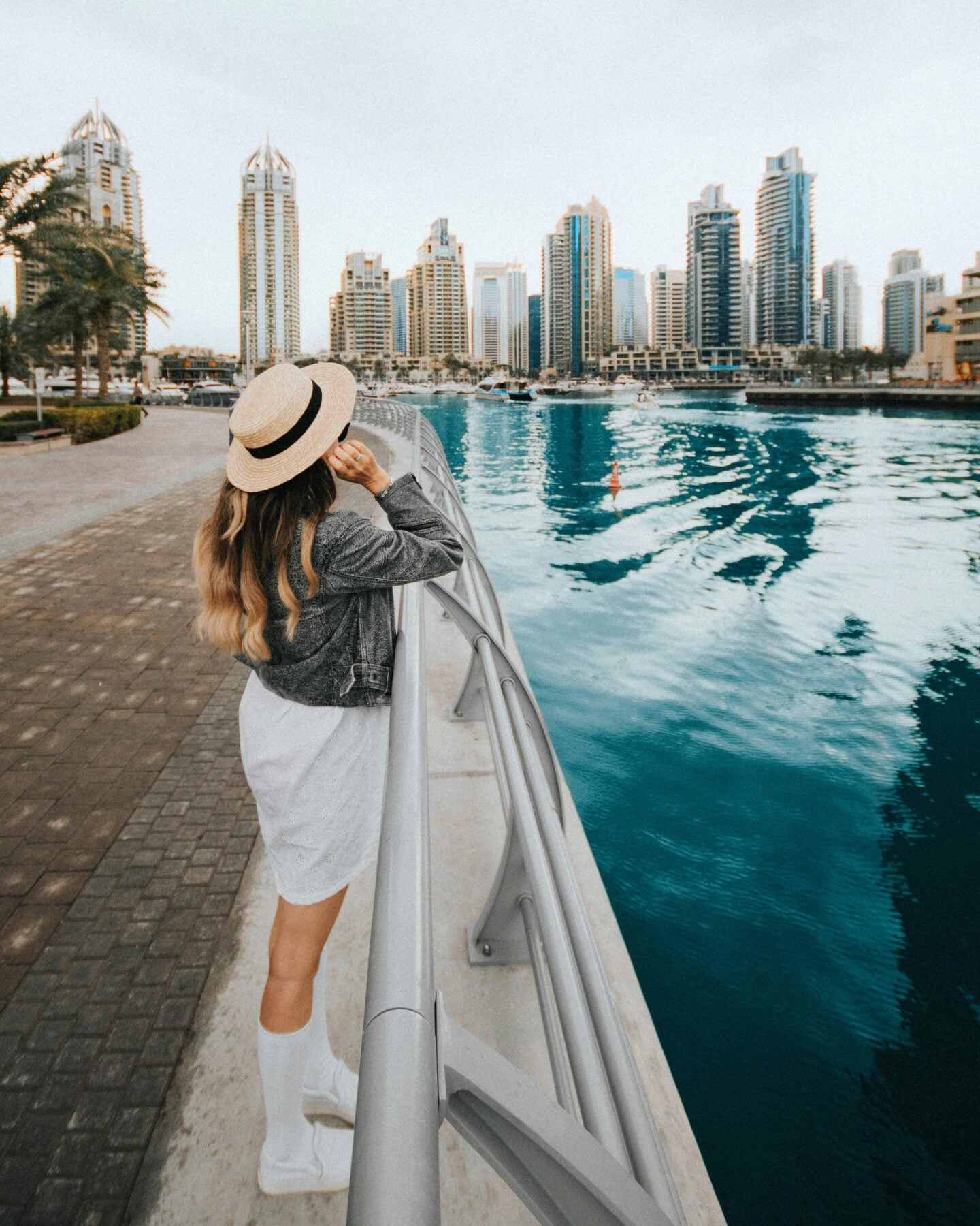 A woman wearing a hat and white dress looks out over the Dubai Marina waterfront with tall skyscrapers in the background.