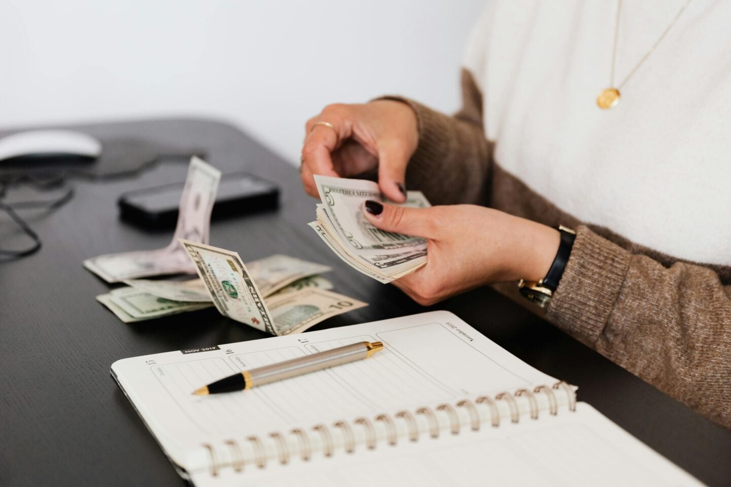 Woman counting US dollar bills at a desk with a planner and smartphone nearby.