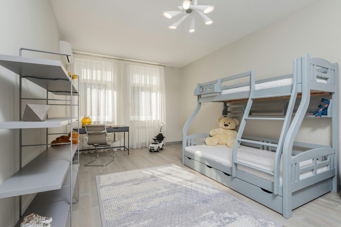 Spacious children’s bedroom with grey bunk beds, a large teddy bear, modern shelving, and a desk by sheer-curtained windows.