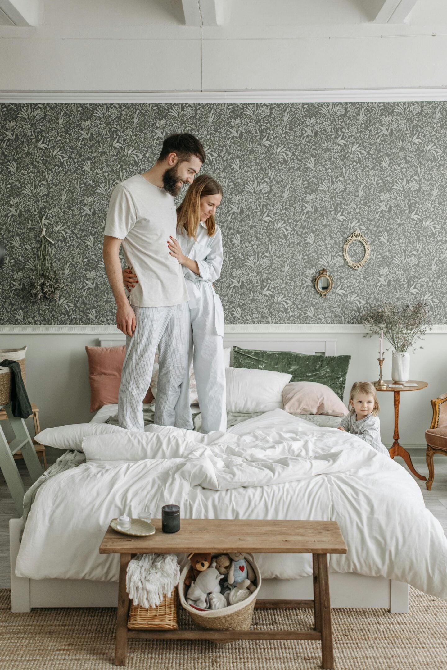 Happy young family with a small child playing on a bed in a stylish bedroom with floral wallpaper and cozy decor.
