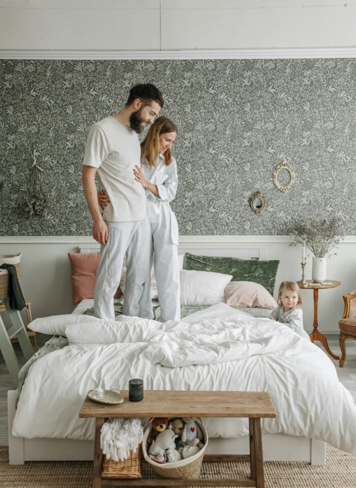 Happy young family with a small child playing on a bed in a stylish bedroom with floral wallpaper and cozy decor.