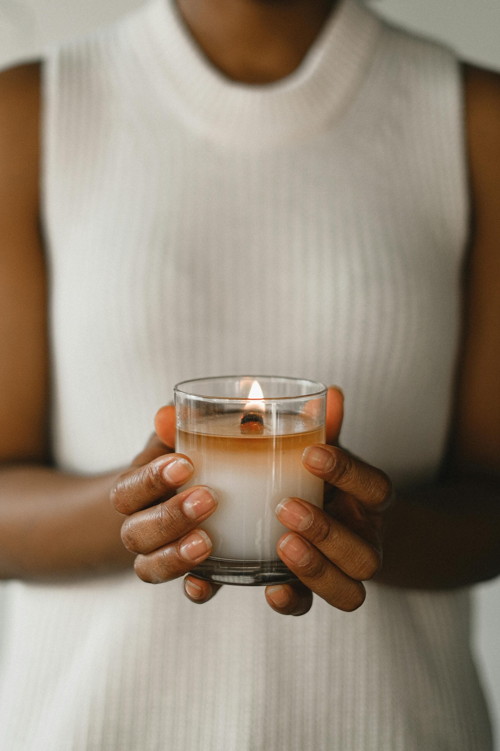 Person in a sleeveless white sweater holding a lit glass candle, creating a calm and cozy atmosphere.