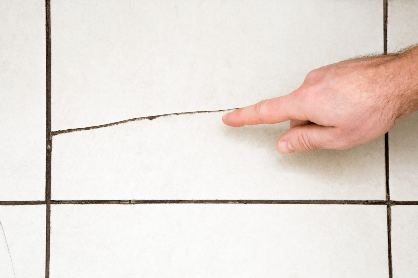 Close-up of a person’s hand pointing at a long crack running across a white ceramic floor tile with dark grout lines.