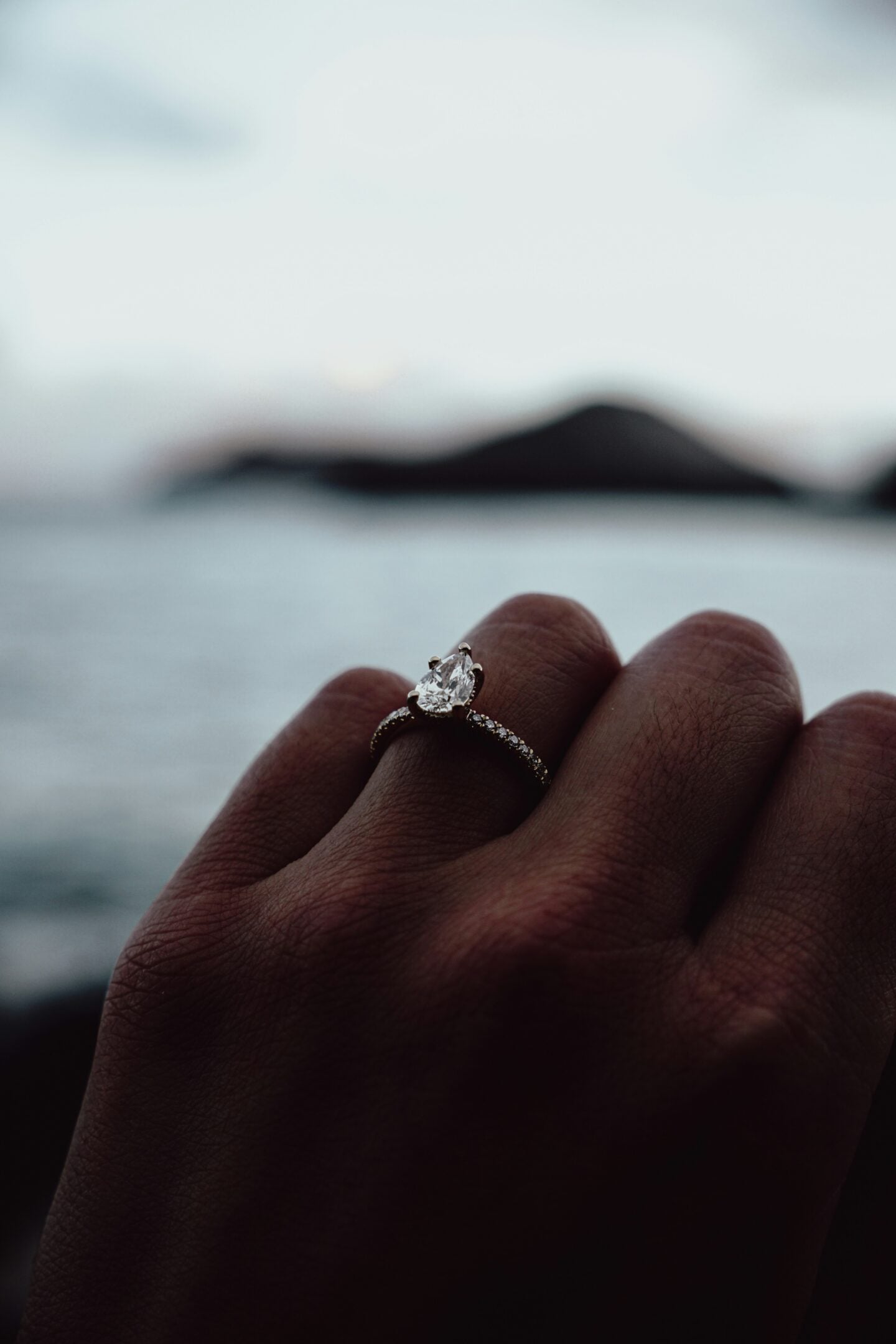 Hand wearing a pear-shaped diamond engagement ring with a delicate band near the ocean at sunset.