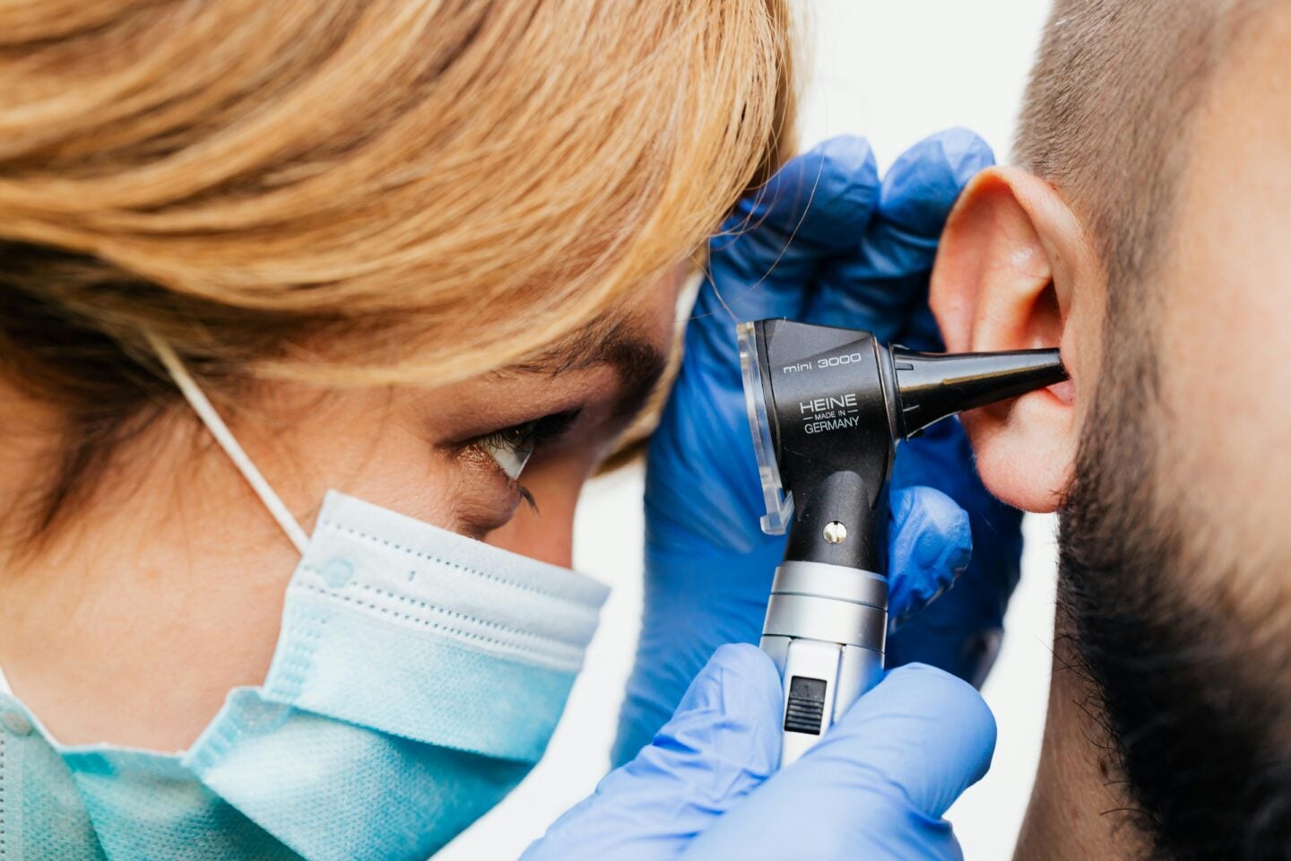 Healthcare professional in a face mask and blue gloves examining a patient’s ear with an otoscope.