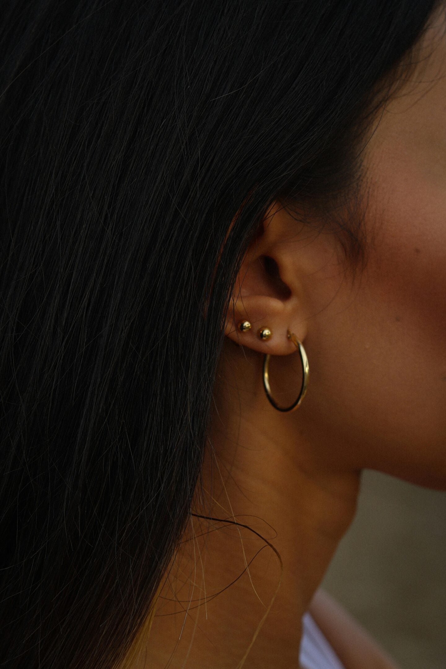 Side view of a woman’s ear with long dark hair, wearing three small gold studs and a gold hoop earring.