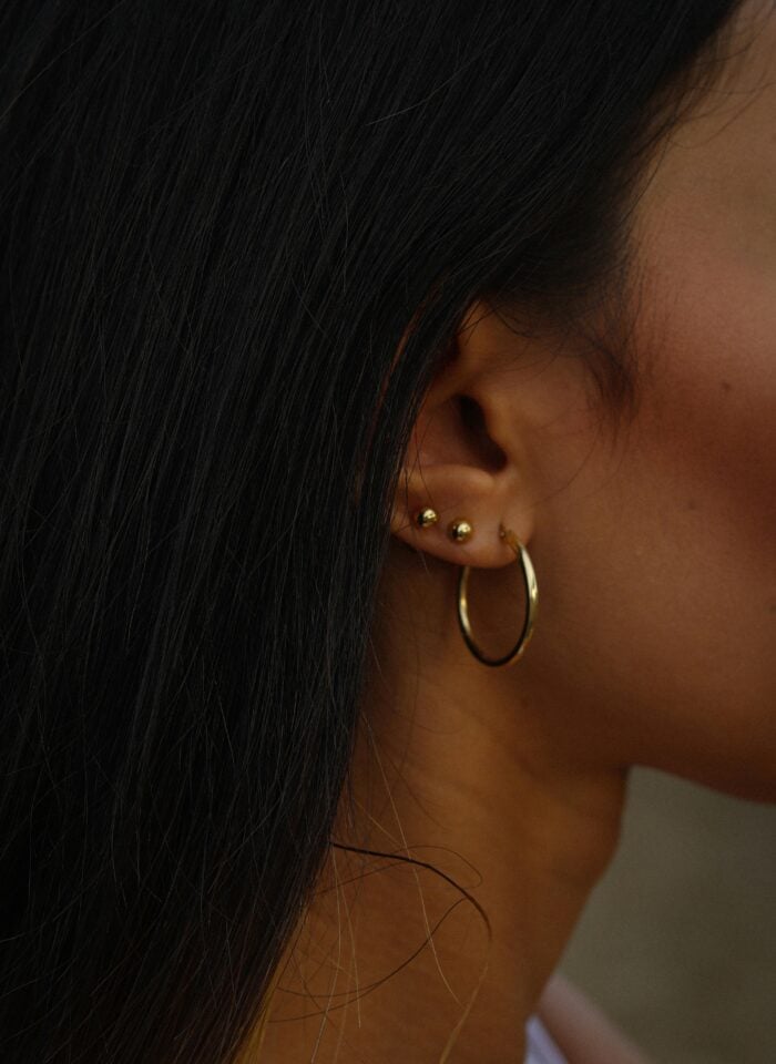 Side view of a woman’s ear with long dark hair, wearing three small gold studs and a gold hoop earring.
