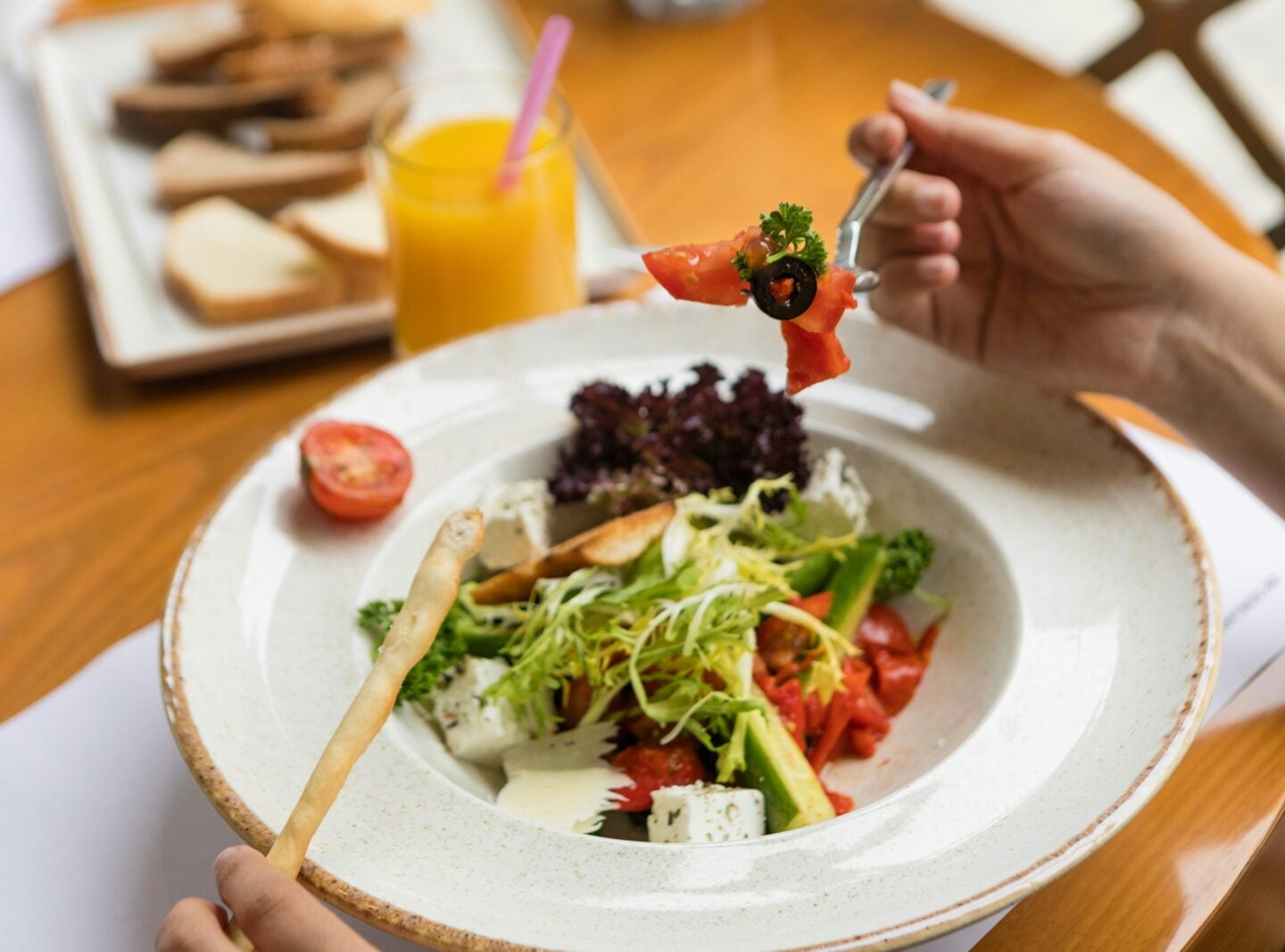 Close-up of a person eating a fresh salad with vegetables, feta cheese, and olives at a dining table, with bread and a glass of orange juice in the background.