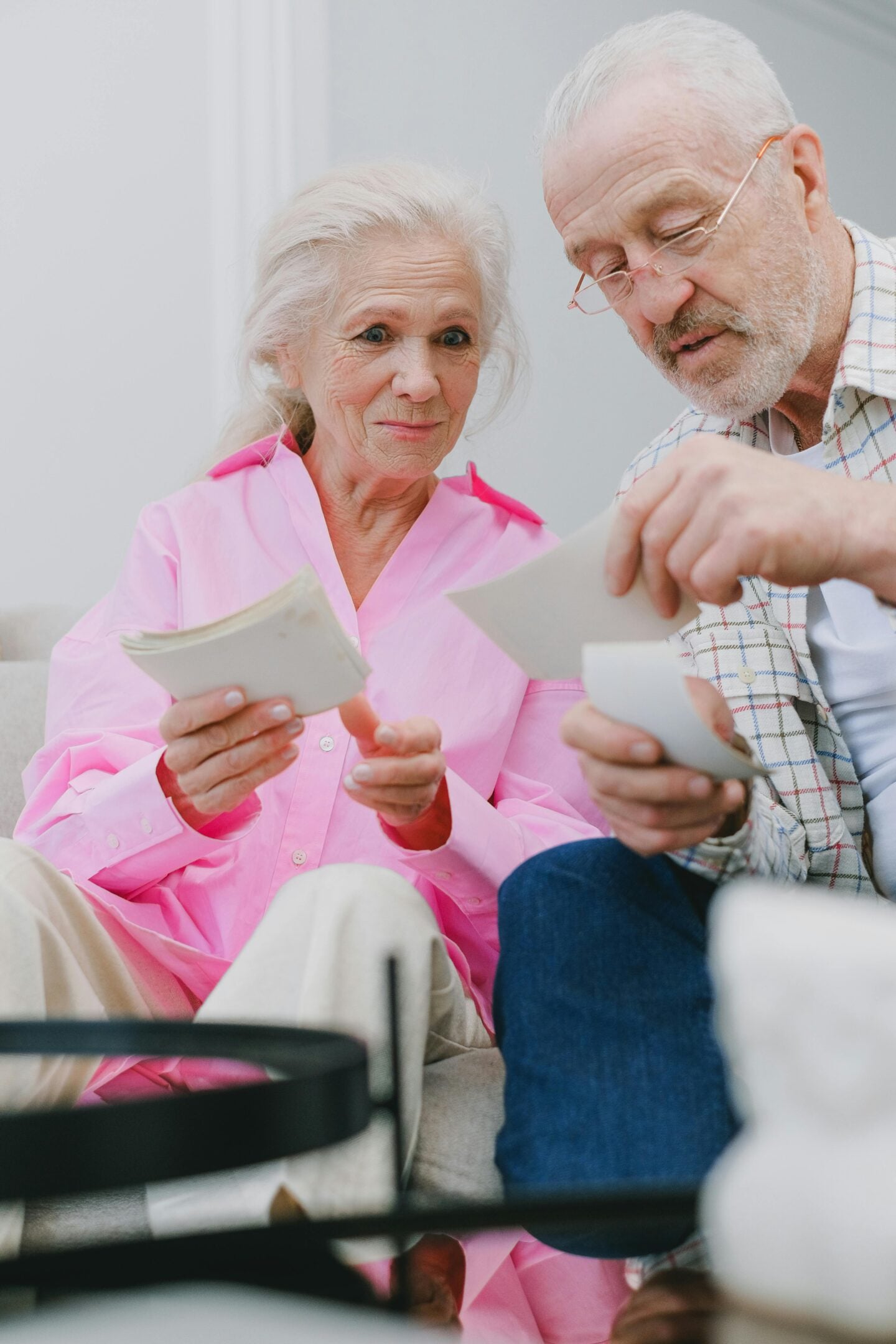 Elderly couple sitting together on a couch, smiling and holding photographs as they reminisce.