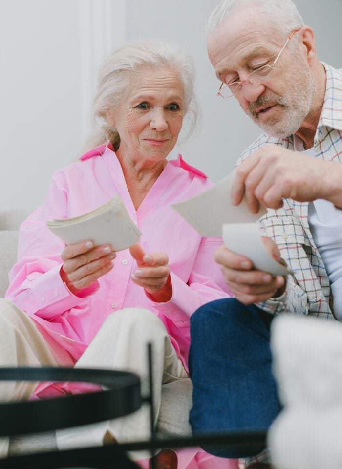 Elderly couple sitting together on a couch, smiling and holding photographs as they reminisce.