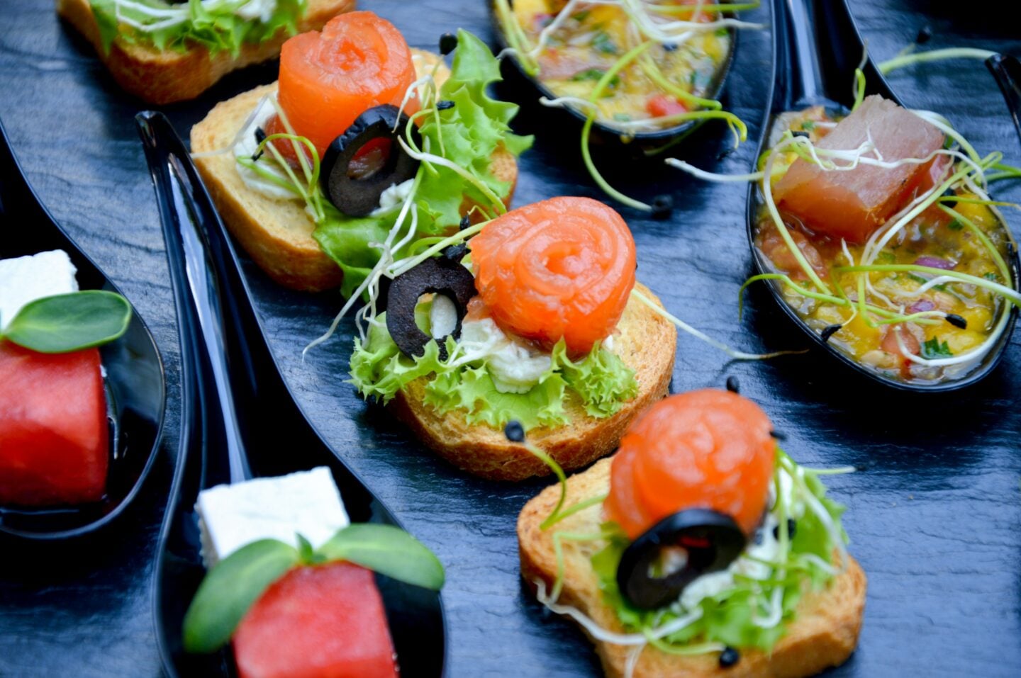 Elegant event appetizers displayed on black serving platters, including salmon and cream cheese bruschetta with olives and greens, watermelon and feta bites, and tuna tartare with sprouts in black spoons.
