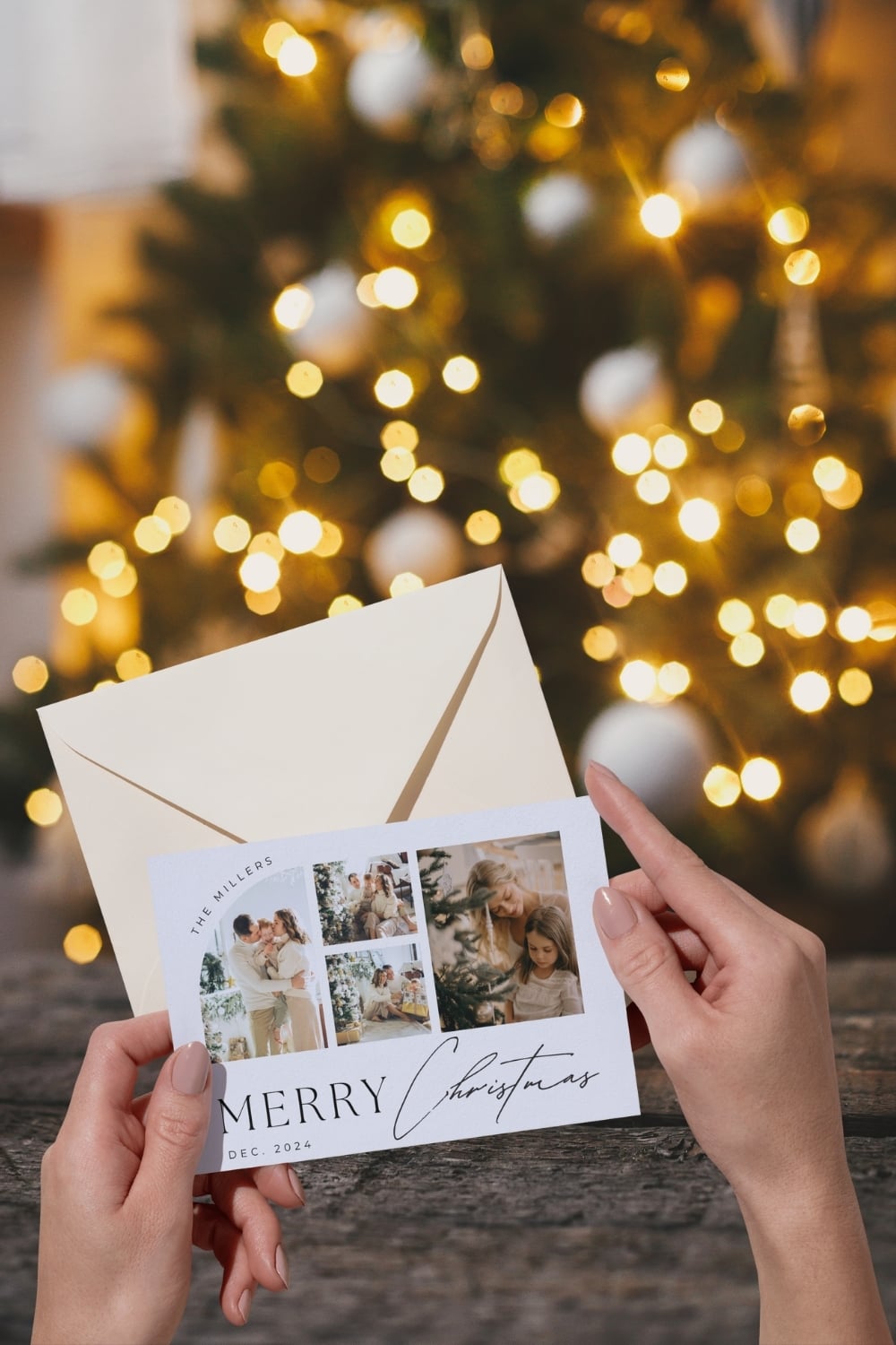 Hands holding a family Christmas card with multiple photos and an envelope in front of a glowing Christmas tree with warm lights.