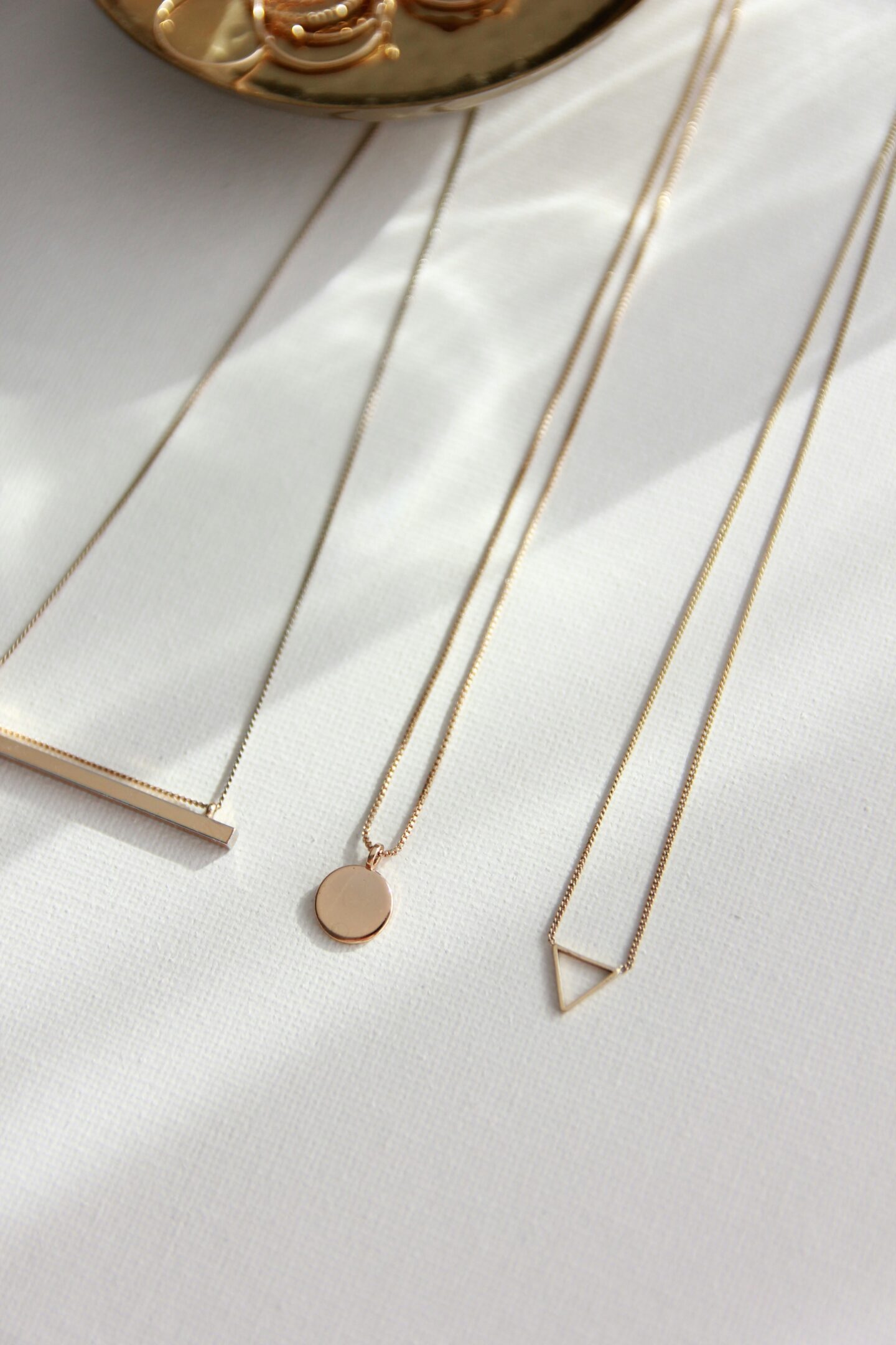 Minimalist gold necklaces with geometric pendants—a bar, a circle, and a triangle—arranged on a white fabric surface next to a small dish of gold rings.