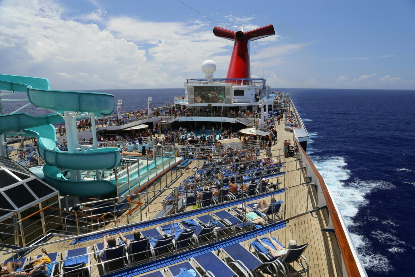 Busy cruise ship deck with sun loungers, swimming pools, and a large waterslide.