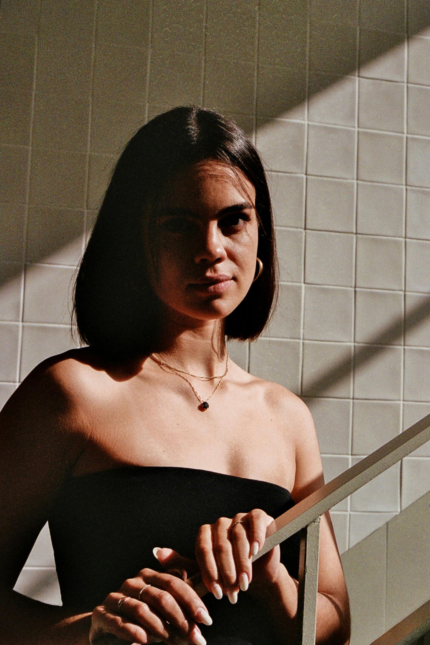 Young woman wearing layered gold necklaces with a small gemstone pendant, standing by a tiled wall in natural light.