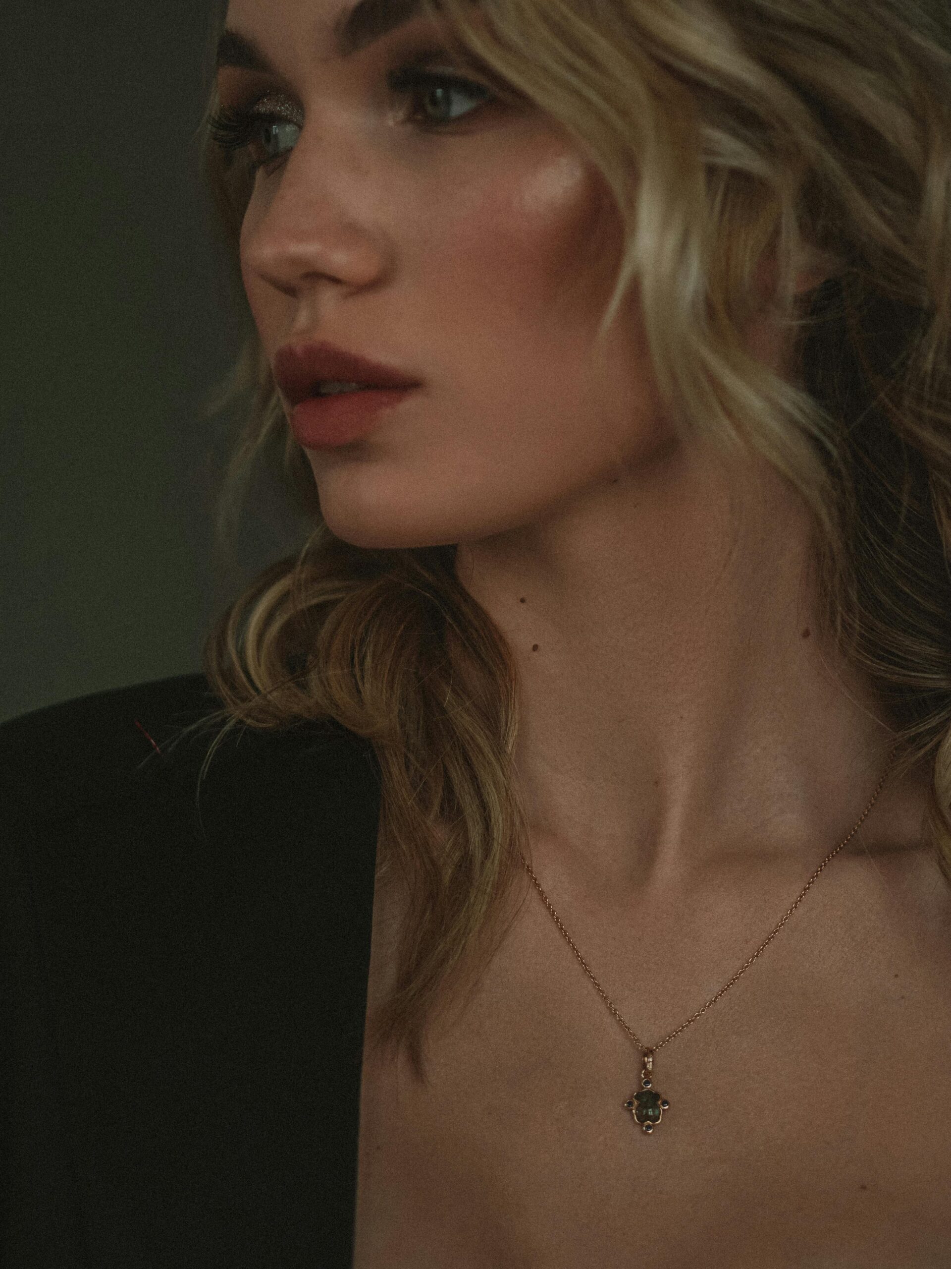 Close-up portrait of a woman in a black blazer wearing an elegant gold chain with a small dark gemstone pendant.