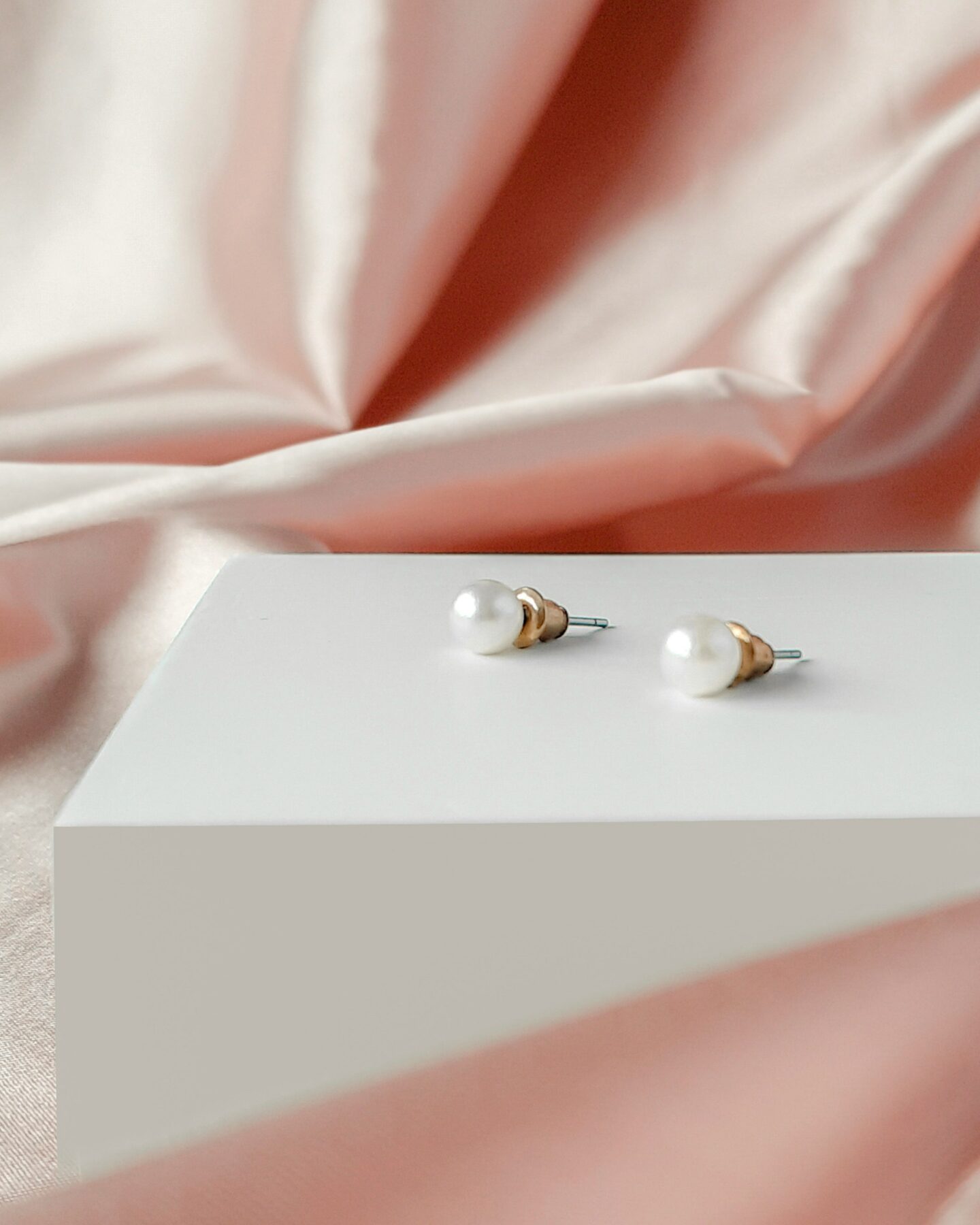 Elegant pair of minimalist pearl stud earrings displayed on a white block with soft pink satin fabric in the background.