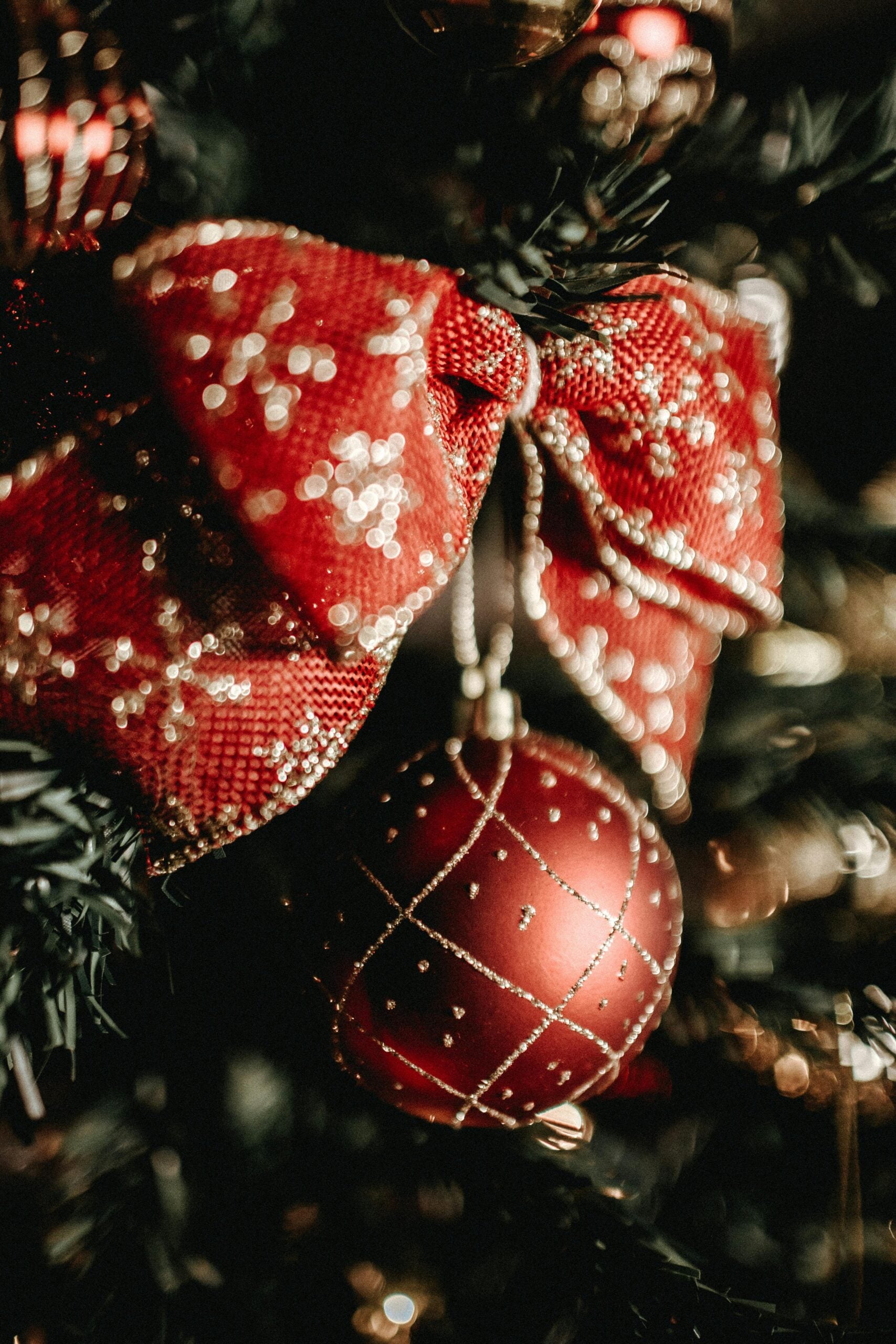 Festive red bow and glittering bauble ornament on a Christmas tree with twinkling lights.
