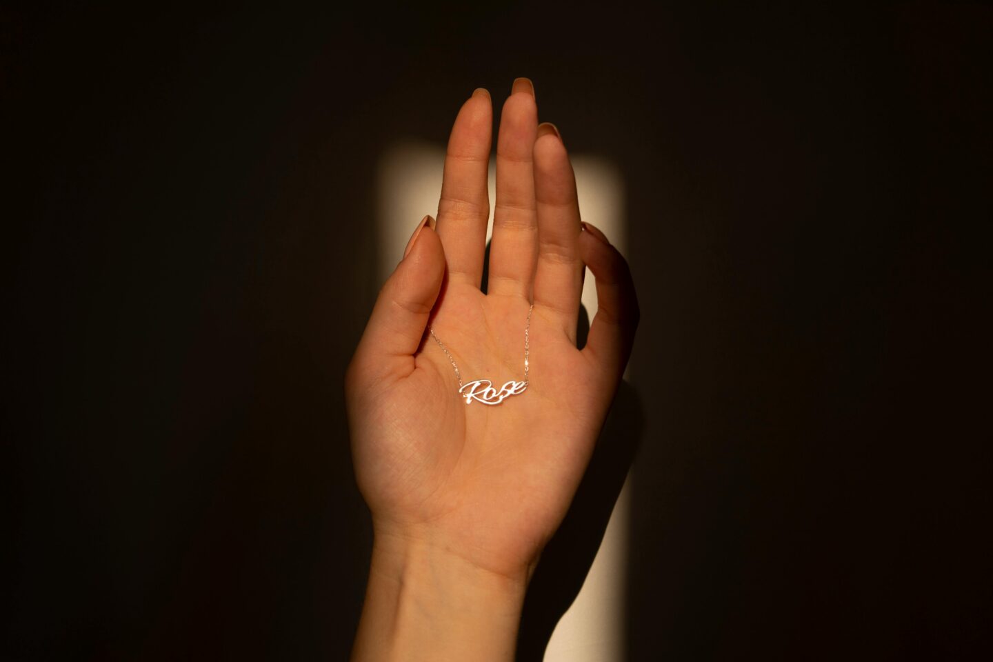 Hand holding a delicate silver necklace with a pendant that spells “Rose,” highlighted by soft sunlight against a dark background.
